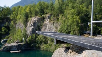 Metallene Cliffdiving-Plattform ragt über Felsen und türkisfarbenes Wasser vor bewaldeten Bergen