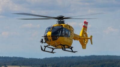 Gelber Rettungshubschrauber mit rot-weißem Heck im Flug über bewaldeter Landschaft