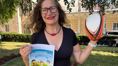 Frau mit Brille und rotem Lippenstift hält Broschüre und Rugbyball in die Kamera