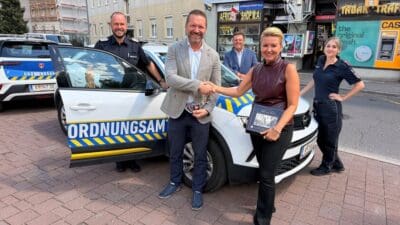 Mann und Frau schütteln sich vor einem weißen Ordnungsamts-Fahrzeug die Hände, weitere Personen im Hintergrund