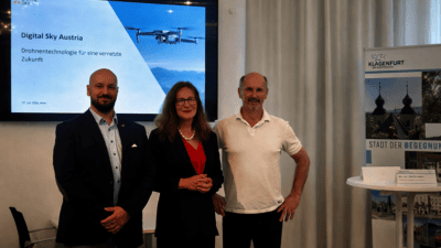 Drei Personen stehen vor einem Bildschirm mit Digital Sky Austria Drohnenpräsentation und Klagenfurt Banner