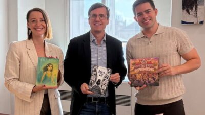 Eine Frau in einer gestreiften Jacke und zwei Männer halten Bücher hoch und lächeln in die Kamera.