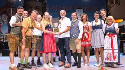 Männer und Frauen in traditioneller Tracht stehen bei der Siegerehrung mit einem Pokal auf der Bühne