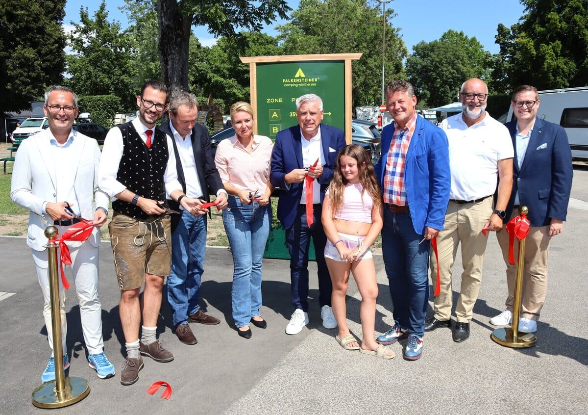 Mehrere Personen in formeller und traditioneller Kleidung bei der Banddurchschneidung vor dem Schild des Falkensteiner Camping Wörthersee.