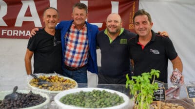 Vier Männer lächeln hinter Schalen mit grünen und schwarzen Oliven auf einem Feststand.