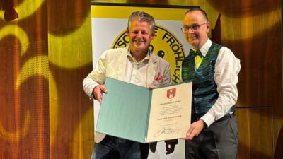 Zwei Männer präsentieren eine geöffnete Mappe mit Urkunde der Musikschule Fröhlich.