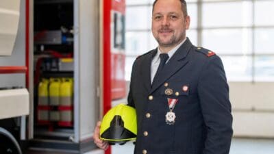 David Walder in dunkelblauer Feuerwehruniform mit Orden und gelbem Helm in der Hand vor einem Einsatzfahrzeug.