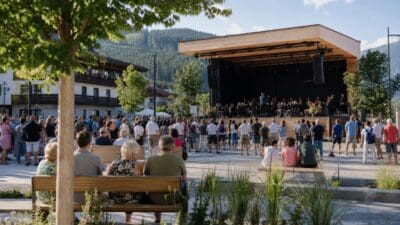 Menschen sitzen auf Bänken und stehen vor einer Bühne mit Orchester im sonnigen Dorfplatz