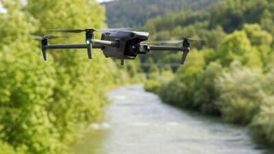 Eine graue Drohne mit vier rotierenden Propellern fliegt über einem Fluss vor grünen Bäumen.