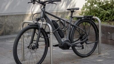 Graues E-Bike mit Kabelschloss an einem silbernen Fahrradständer auf Pflastersteinen