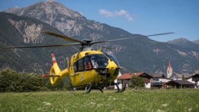 Gelber Rettungshubschrauber mit laufendem Rotor auf Wiese vor Bergmassiv und Dorf mit Kirchturm