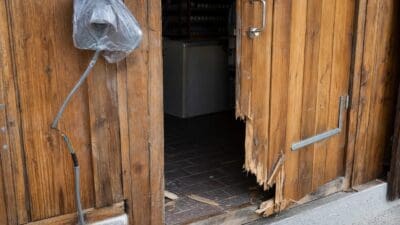 Aufgebrochene Holztür eines Ladens mit in Plastik verhüllter Überwachungskamera und freiliegenden Kabeln