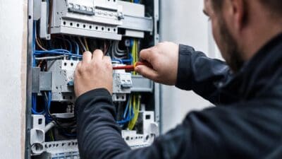 Elektriker arbeitet mit einem Schraubendreher an Schaltern eines geöffneten Verteilerkastens mit bunten Kabeln