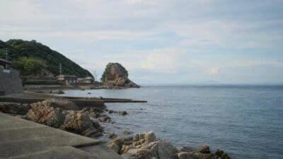 Felsige Küste mit Betonweg, Häusern am grünen Hang und einem Felsen im Meer bei Kumamoto