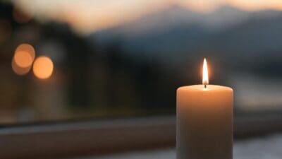 Eine brennende weiße Kerze vor unscharfen Lichtern und Bergen bei Dämmerung.