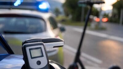 Nahaufnahme eines Alkoholtestgeräts vor einem Polizeiauto mit Blaulicht und einem E-Scooter am Straßenrand