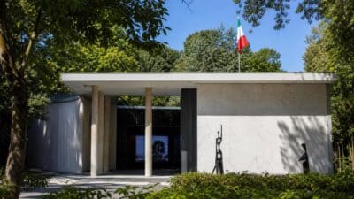 Weißer moderner Pavillon mit Säulen, italienischer Flagge und abstrakten Skulpturen vor Bäumen in Venedig.