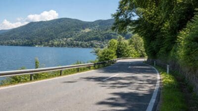 Geschwungene Asphaltstraße am Ufer eines großen Sees mit grünen Hügeln und Bäumen