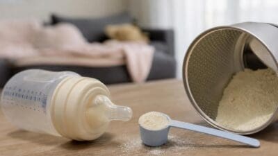 Transparente Babyflasche mit Milch, blauer Messlöffel und offene Dose Babynahrung auf Holztisch.