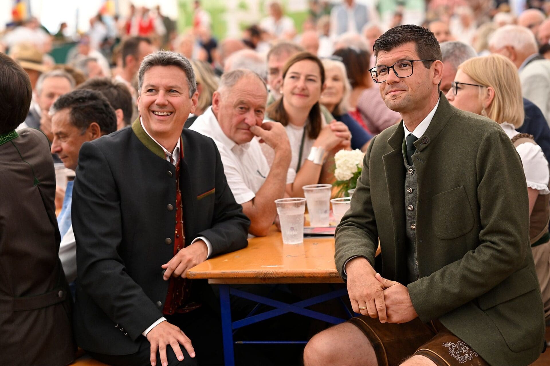 Zwei Männer in Tracht sitzen an einem Holztisch mit Bechern vor Publikum.