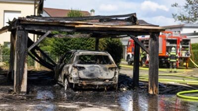 Ausgebranntes Auto unter verkohltem Carport mit Feuerwehrfahrzeug im Hintergrund