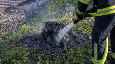Feuerwehrmann in Schutzkleidung sprüht Wasser auf brennenden Baumstumpf an der Bahntrasse