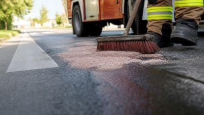Feuerwehrmann in Schutzkleidung mit gelben Reflexstreifen kehrt rosa Absorptionsgranulat auf asphaltierten Straße vor Feuerwehrfahrzeug