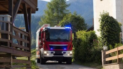 Rotes Feuerwehrfahrzeug mit blauem Blaulicht fährt auf einer Straße zwischen Häusern und Bergen.