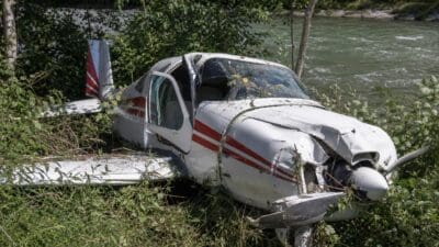 Ein stark beschädigtes weiß-rotes Flugzeug liegt im dichten Gebüsch am Flussufer.