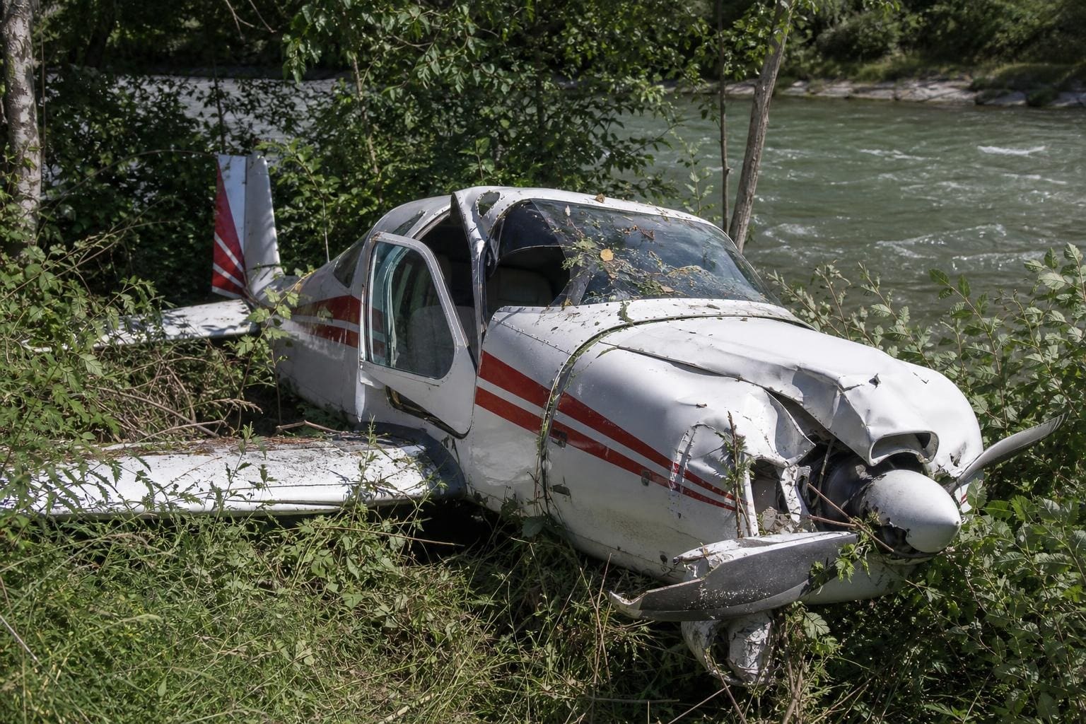 Ein stark beschädigtes weiß-rotes Flugzeug liegt im dichten Gebüsch am Flussufer.