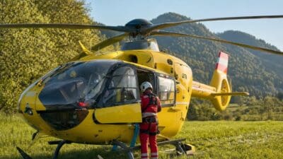 Gelber Rettungshubschrauber mit geöffneter Tür auf einer Wiese, Rettungskraft in roter Uniform steht daneben