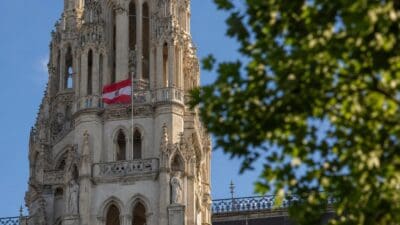 Reich verzierter gotischer Turm mit österreichischer Flagge und Statuen vor blauem Himmel, grünes Laub im Vordergrund