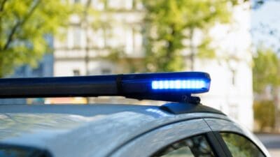 Blaues Blaulicht leuchtet auf dem Dach eines Polizeifahrzeugs vor unscharfen Häusern und Bäumen.