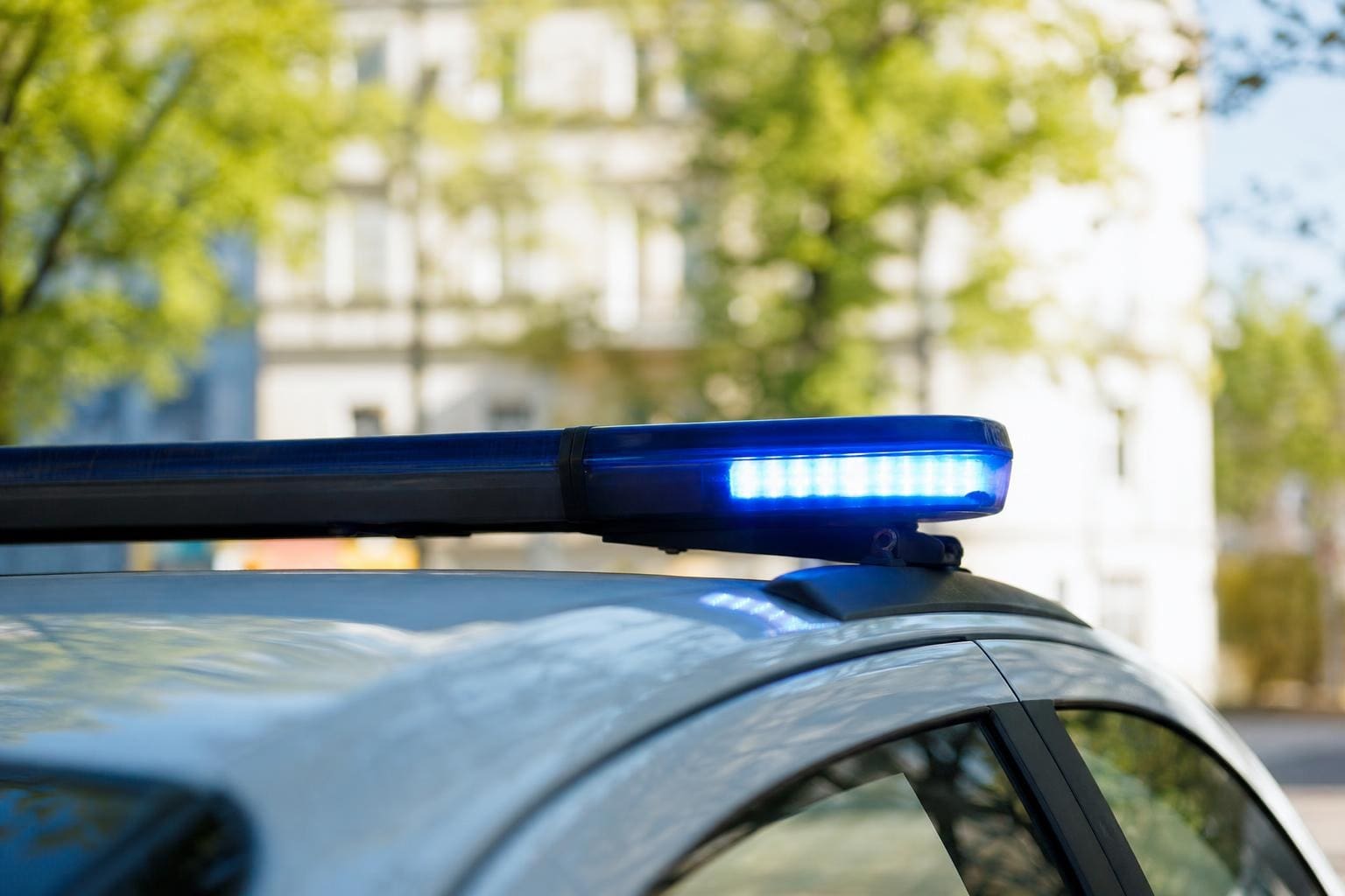 Blaues Blaulicht leuchtet auf dem Dach eines Polizeifahrzeugs vor unscharfen Häusern und Bäumen.
