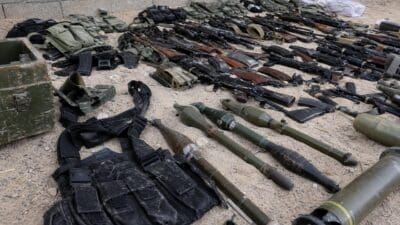 Zahlreiche Gewehre, Raketenwerfer und Munition liegen auf sandigem Boden ausgebreitet.