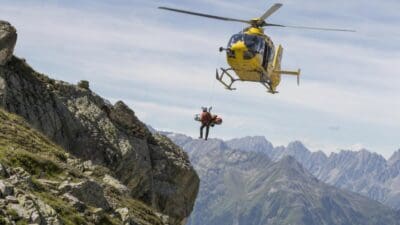 Gelber Rettungshubschrauber mit Bergretter an Seil über felsigem Hochgebirge