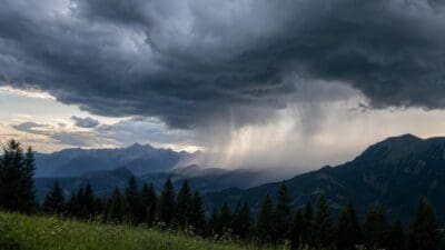 Dunkle Gewitterwolken mit herabfallendem Regen über bewaldeten Bergen und grüner Wiese