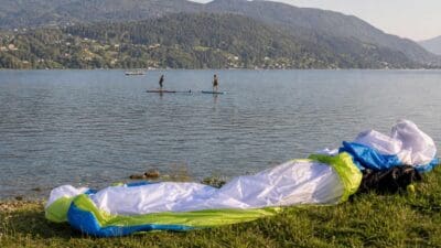 Ein bunter Gleitschirm liegt am grasigen Ufer des Ossiacher Sees mit zwei Stand-up-Paddlern auf dem Wasser im Hintergrund.