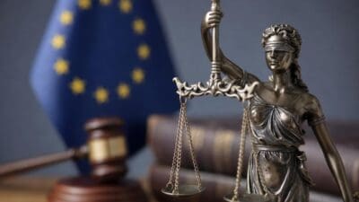Bronzestatue der Justitia mit Waage und Schwert vor der EU-Flagge und einem Richterhammer