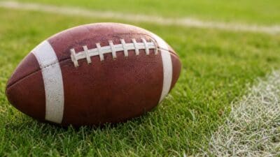 Ein brauner American Football mit weißen Streifen liegt auf grünem Rasen.