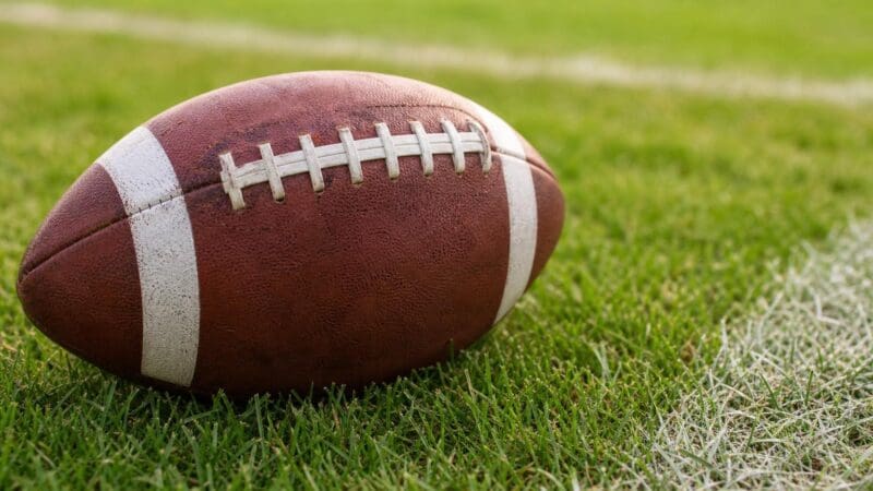 Ein brauner American Football mit weißen Streifen liegt auf grünem Rasen.