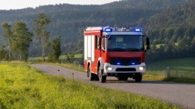 Rotes Feuerwehrfahrzeug mit blauen Blinklichtern fährt auf einer Landstraße vor Hügeln.