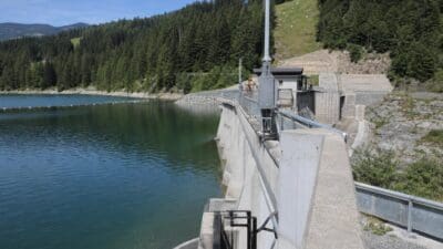 Betonstaumauer eines Rückhaltebeckens mit grünem Wasser, Nadelwäldern und Bergen unter blauem Himmel