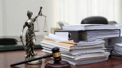 Justitia-Statue mit Waage und Schwert neben Richterhammer und Aktenstapeln auf einem Holztisch