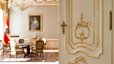 Weiße Tür mit aufwendigen goldenen Verzierungen vor einem historischen Raum mit Kronleuchter