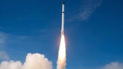 Weiße Rakete mit schwarzer Spitze steigt mit hellem Flammenschweif in den blauen Himmel