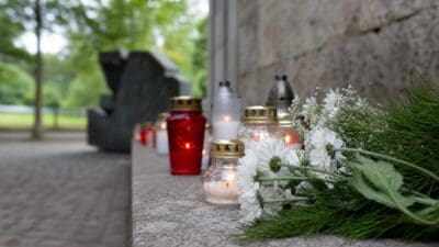 Brennende Kerzen in roten und durchsichtigen Gläsern neben weißen Blumen auf einem Steinabsatz an einer Gedenkstätte.