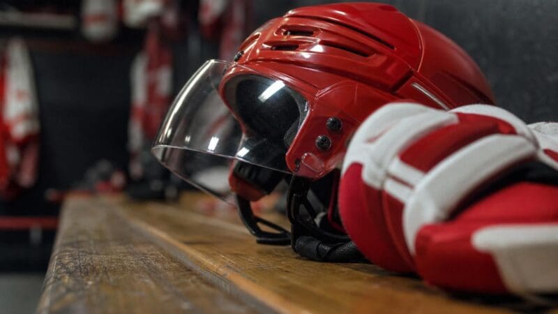 Roter Eishockeyhelm mit Visier und rote weiße Handschuhe auf einer Holzbank