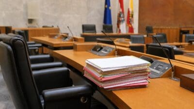 Stapel bunter Aktenordner auf einem Holztisch im Plenarsaal mit Fahnen im Hintergrund