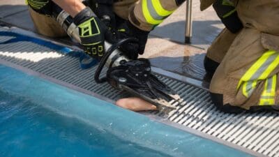 Feuerwehrleute in Schutzkleidung setzen einen Spreizer am Fuß eines Kindes an einem Metallgitter im Schwimmbecken ein